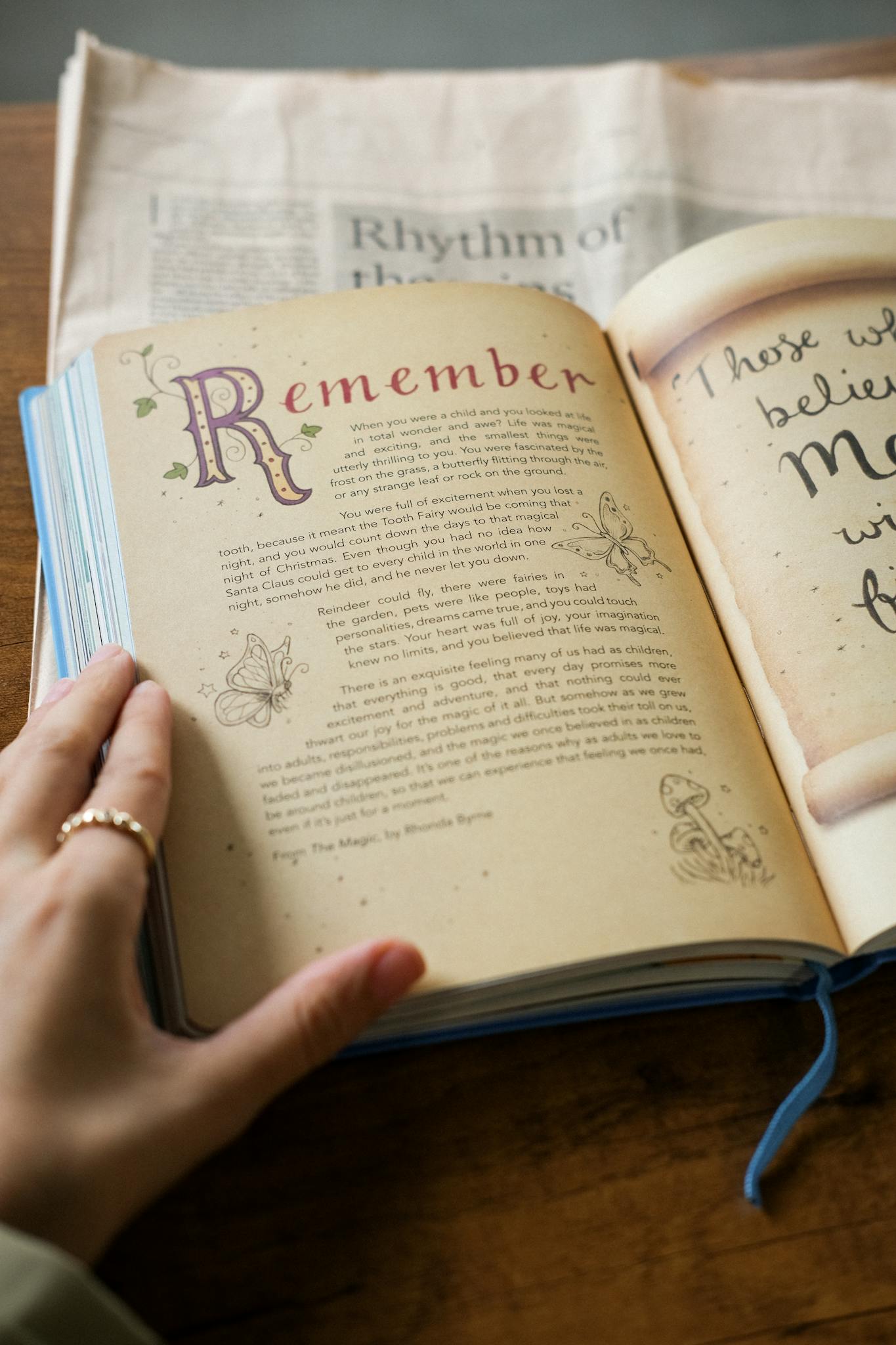 An open book with decorative text and hand, placed on a wooden table with a newspaper backdrop.