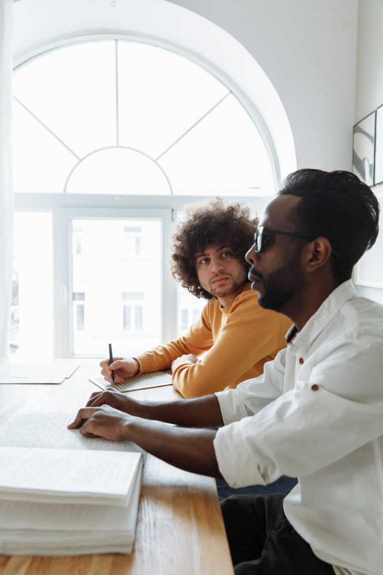 Two men engaging in a Braille reading session at a wooden table under natural light.
