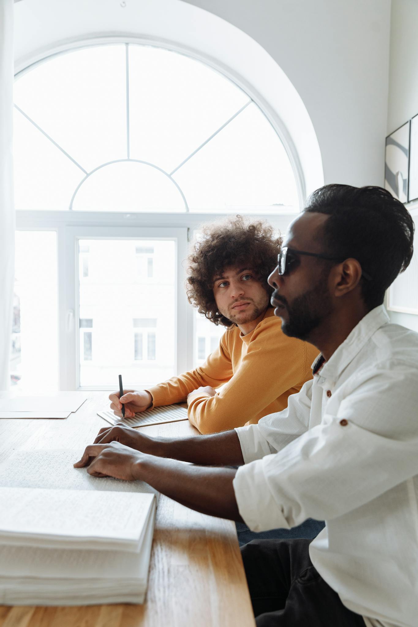 Two men engaging in a Braille reading session at a wooden table under natural light.